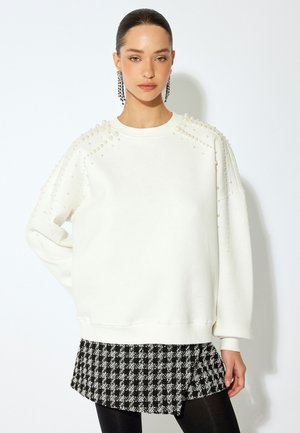 Witte sweatshirt met parelaccenten op de schouders en lange mouwen, gelaagd over een zwart-wit geruite rok, gecombineerd met zwarte leggings.