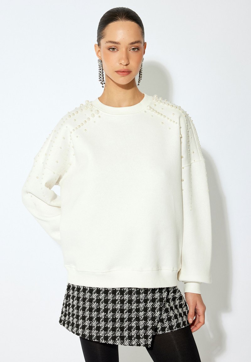 Sudadera blanca con adornos de perlas en los hombros y mangas largas, combinada con una falda de cuadros en blanco y negro, y leggings negros.