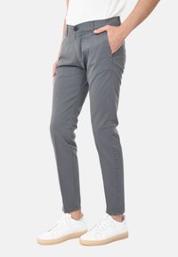 Pantaloni grigi slim-fit con una texture liscia, dotati di tasche laterali, zip e chiusura con un solo bottone, abbinati a sneakers bianche.