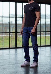 Czarna koszulka z krótkim rękawem z graficznym detalem, niebieskie dżinsy oraz białe trampki. Tło zawiera duże okna i przemysłową podłogę.