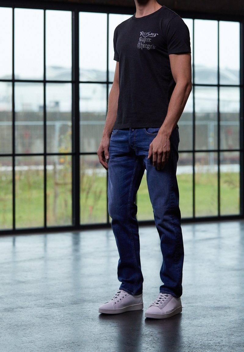 Czarna koszulka z krótkim rękawem z graficznym detalem, niebieskie dżinsy oraz białe trampki. Tło zawiera duże okna i przemysłową podłogę.
