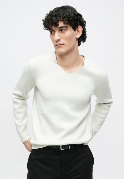 Λευκό πουλόβερ με λαιμόκοψη σε σχήμα V, κατασκευασμένο από απαλό, υφασμένο υλικό. Μακριά μανίκια, άνετη γραμμή, συνδυασμένο με μαύρο παντελόνι και ζώνη.