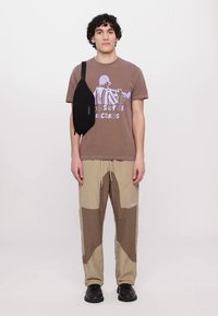 Brun grafisk t-shirt med lilla print, parret med beige og brune patchwork-bukser; sort crossbody taske; sorte sko; neutral baggrund.