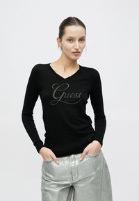 Jersey negro de manga larga con cuello en V y logo plateado de "Guess", con puños y dobladillo acanalados. Combina con pantalones plateados brillantes.