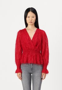 Maglione rosso peloso con maniche lunghe, scollo a V profondo e vita arricciata. Presenta una finitura testurizzata e polsini a campana. Abbinato a jeans grigi.