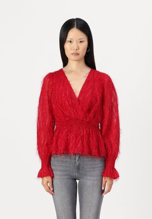 Pull en laine rouge moelleux avec des manches longues, décolleté en V profond et taille smockée. Présente une finition texturée et des poignets en cloche. Associé à un jean gris.