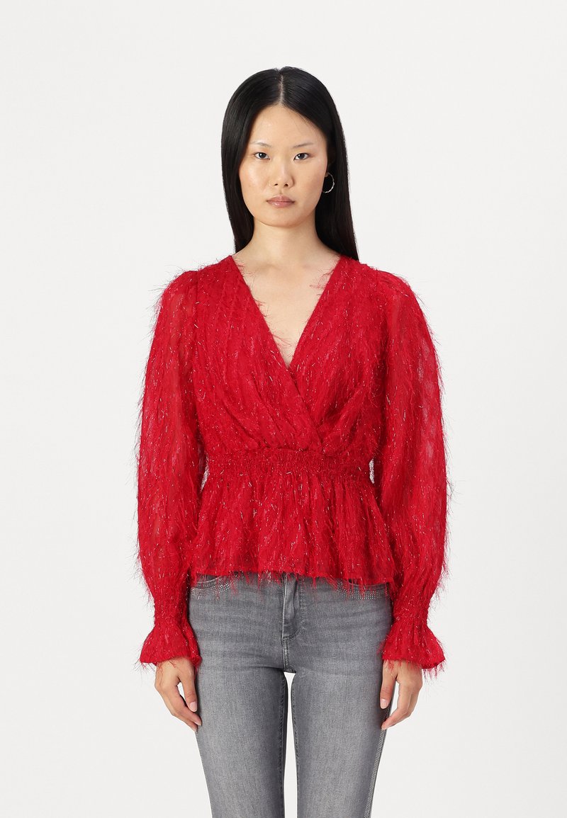 Maglione rosso peloso con maniche lunghe, scollo a V profondo e vita arricciata. Presenta una finitura testurizzata e polsini a campana. Abbinato a jeans grigi.