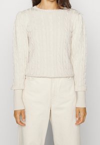 Krémový svetr s copánkovým vzorem, kulatým výstřihem, dlouhými rukávy a přiléhavými žebrovanými manžetami. Kombinován se světlými džínami.