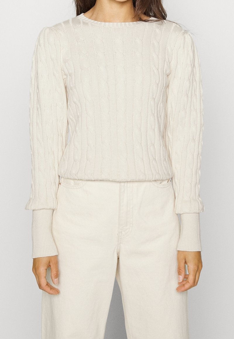 Krémový svetr s copánkovým vzorem, kulatým výstřihem, dlouhými rukávy a přiléhavými žebrovanými manžetami. Kombinován se světlými džínami.
