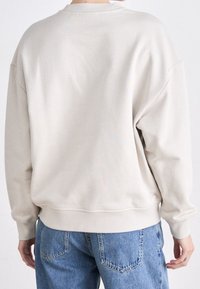 Jasnoszara bluza z okrągłym dekoltem i opuszczonymi ramionami, z prążkowanymi mankietami i dołem, miękka w dotyku, zestawiona z niebieskimi dżinsami.