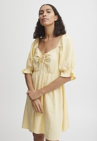 Rochie galbenă din lenjerie, cu decolteu pătrat, mâneci bufante și detalii cu șnur la bust. Siluetă simplă și fluidă, cu o croială casual.