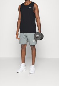 Débardeur sans manches noir Nike et shorts gris clair, tenant un haltère noir. Chaussures de sport blanches et chaussettes montantes avec le logo Nike visible.