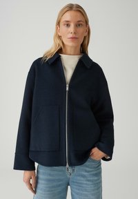 Giacca navy in lana con zip, colletto classico, due tasche frontali, vestibilità rilassata e tessuto morbido. Abbinata a jeans azzurri.