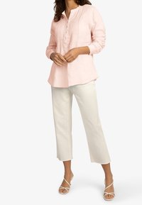Chemise en lin rose clair avec une patte de boutonnage et des plis verticaux, associée à un pantalon blanc cropped, portée avec des talons nud.