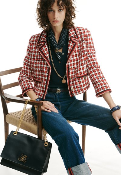 Frau trägt eine rot-weiß karierte Jacke, ein dunkles Hemd mit gepunktetem Schal, blaue Jeans und sitzt auf einem Holzstuhl, hält eine schwarze Handtasche mit Goldkette.