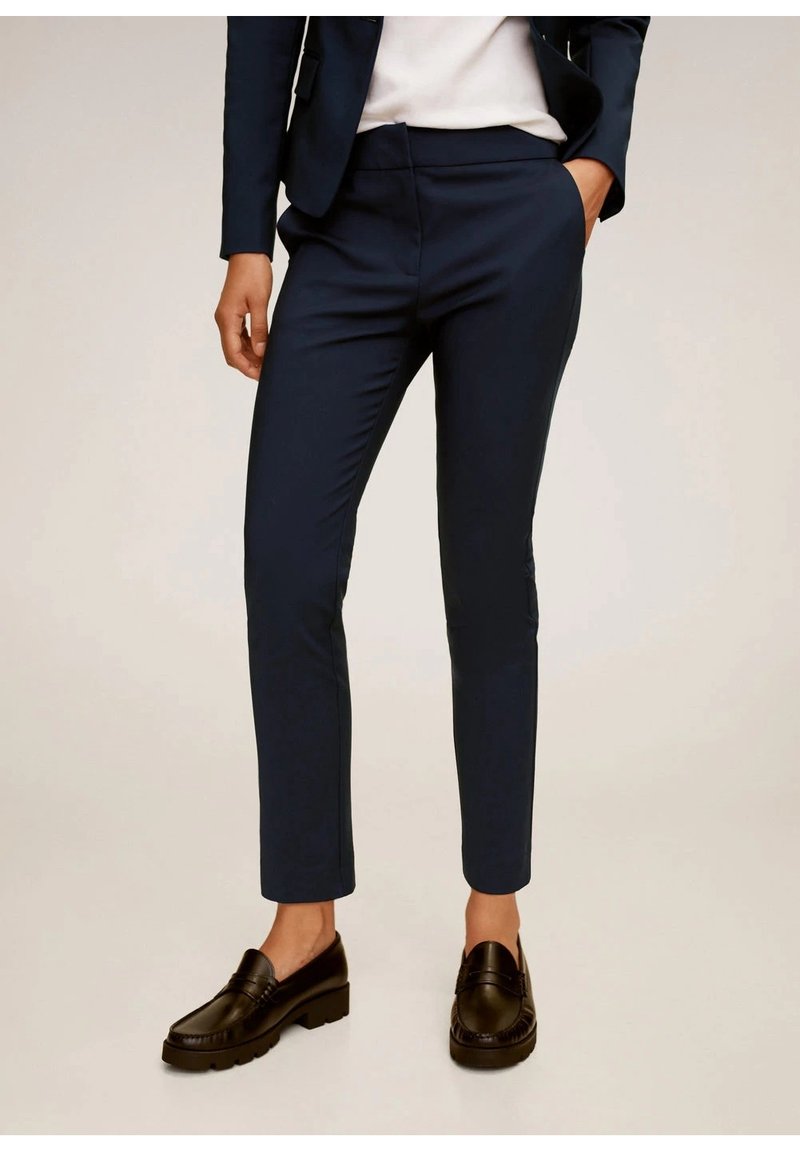 Personne portant un pantalon bleu marine ajusté, un blazer assorti, une chemise blanche et des mocassins noirs, debout avec une main dans la poche. Fond neutre.