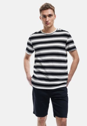 Giovane con i capelli corti che indossa una camicia a righe bianche e nere con maniche corte e pantaloni corti neri, in piedi con le mani in tasca.