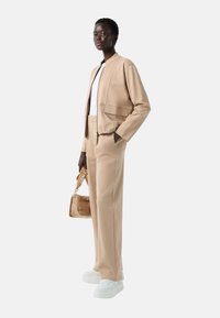 Beige bomberjakke med lynlås og sidelommer, parret med beige bredbenede bukser. Hvide sneakers og en lille tan håndtaske er inkluderet.