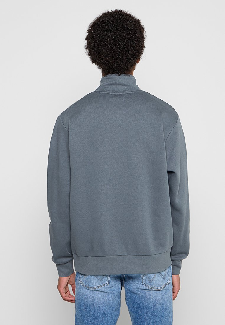 Grauer Fleece-Sweatshirt mit hohem Kragen, gerippten Bündchen und Saum. Hat eine glatte Textur und eine lockere Passform, getragen über blauen Jeans.