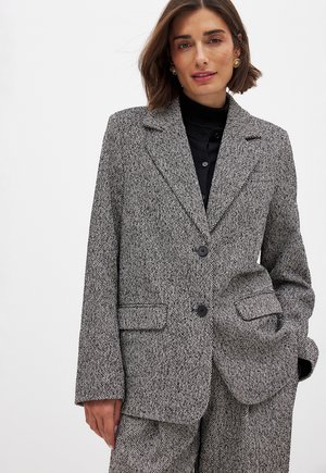 Blazer en pied-de-poule gris avec une coupe ajustée, revers crantés, deux boutons et poches à rabat. Tissu texturé avec une silhouette structurée.
