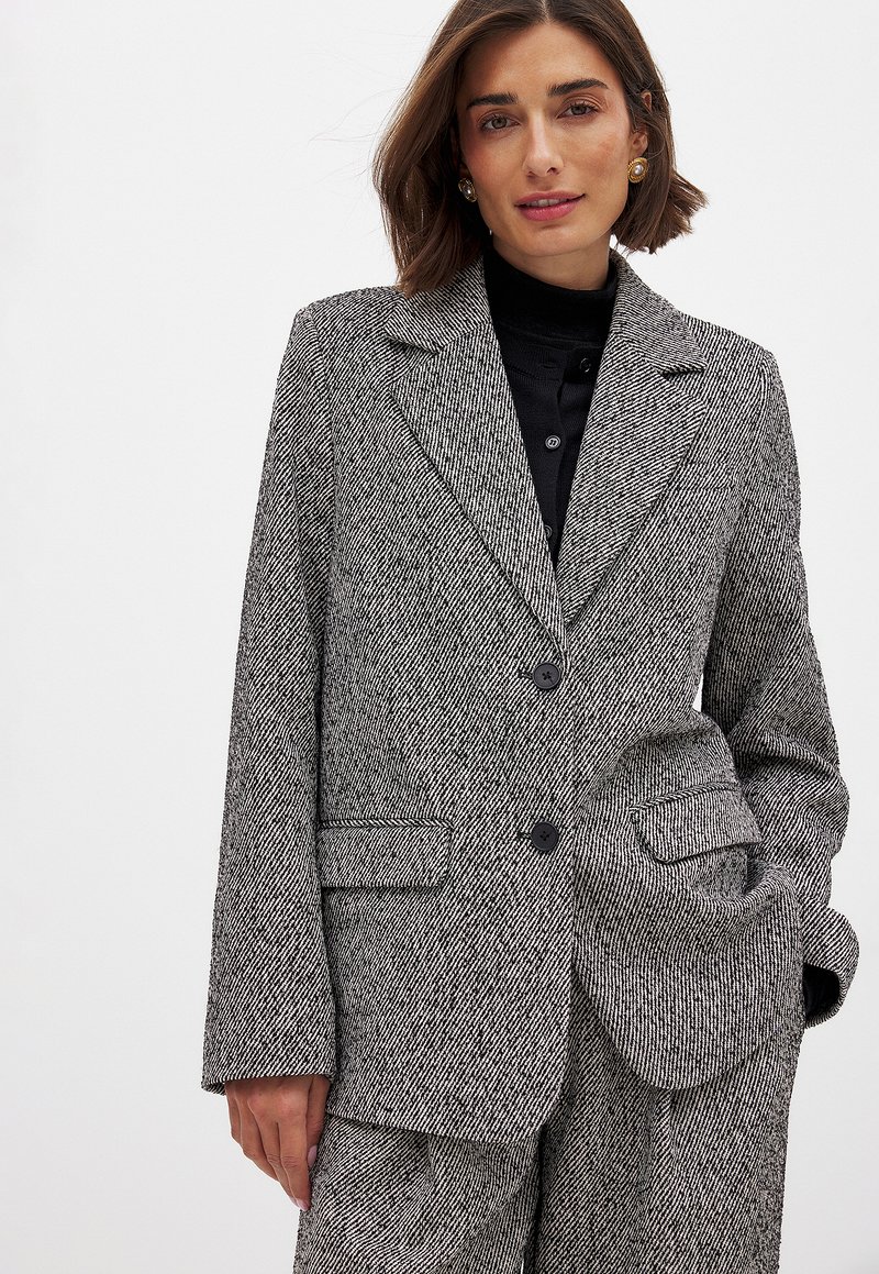 Sivý károvaný blazer s priliehavým strihom, klopovými goliermi, dvoma gombíkmi a záklopkovými vreckami. Textúrovaná látka so štruktúrovanou siluetou.