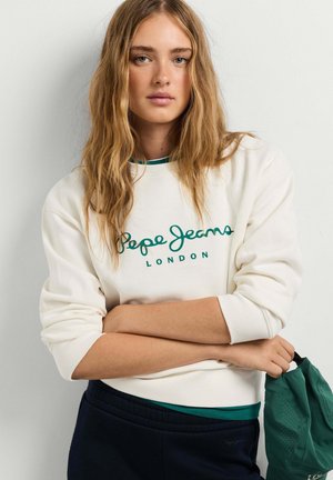 Jonge vrouw met lang blond haar, die een witte Pepe Jeans London sweater draagt, donkere broek en een groene tas vasthoudt met gekruiste armen.