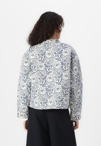 Veste matelassée avec un motif paisley bleu et blanc, col rond, manches longues, silhouette courte et tissu texturé.
