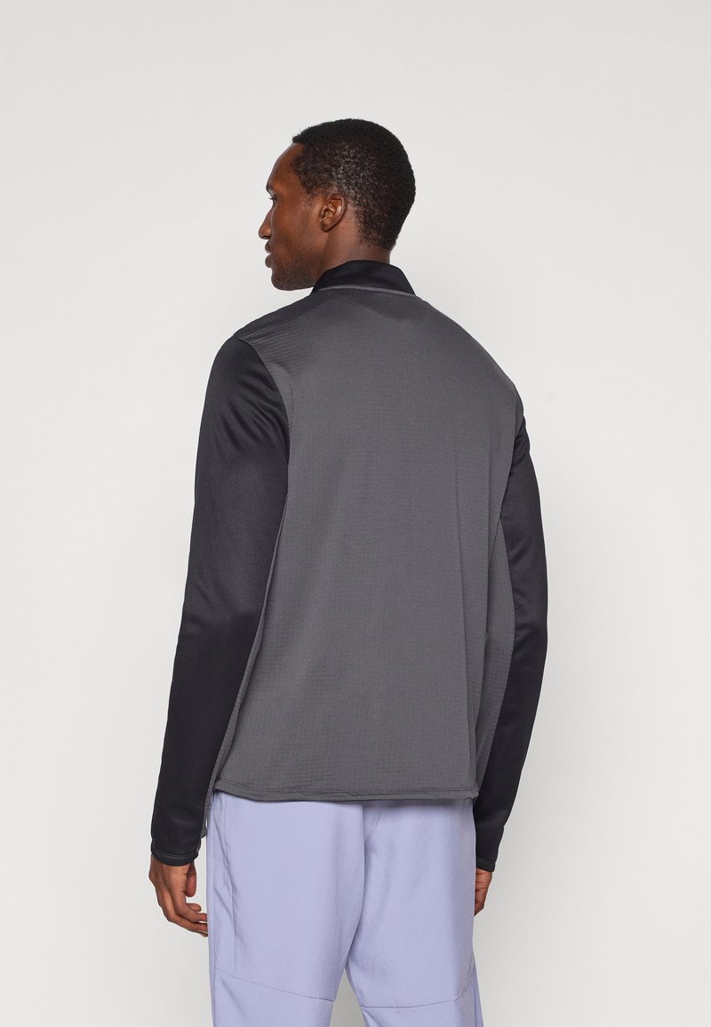 Grå og sort zip-up jakke med lange ærmer, krave og tekstureret stof på ryggen. Har en glat sort accent på ærmerne.