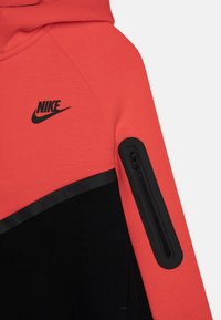 Nike hettegenser med en korallfarge øverste del, svart nedre del, en glidelåslomme på ermet, og en fremtredende svart logo.