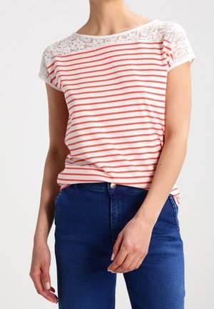 Vrouw die een wit met rood gestreept shirt met korte mouwen en kanten schouders draagt, gecombineerd met een blauwe high-waisted jeans, staand tegen een effen achtergrond.