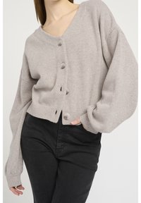 Ljust grå beskuren cardigan med V-ringning, fem stora knappar, sänkt axel och ribbade ärmslut. Bärs över svarta jeans.