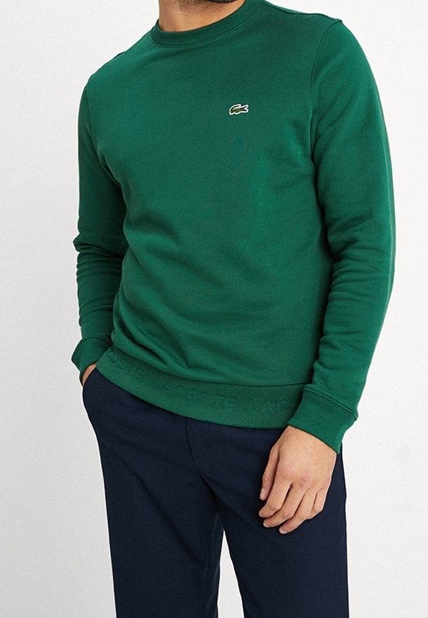 Homme portant un sweat-shirt vert Lacoste à manches longues avec un pantalon bleu marine, debout avec une main dans la poche devant un fond blanc.