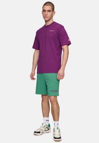 Lilla bomulds t-shirt med korte ærmer, parret med grønne bomulds shorts med "R.S.V.P." broderet i lilla. Sko i blandede farver.
