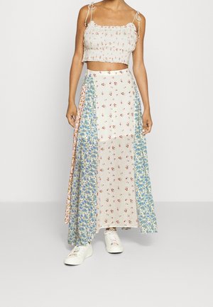 Femme portant un haut court blanc à fines bretelles et une longue jupe patchwork fleurie, associée à des baskets blanches.