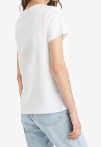 Donna che indossa una semplice maglietta bianca a maniche corte e jeans in denim blu chiaro, in piedi con la schiena parzialmente girata.