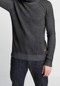 Svart och grå randig tröja med en ribbad textur, hög hals-design och en orange accenttagg i fållen. Långa ärmar med åtsittande mudd.