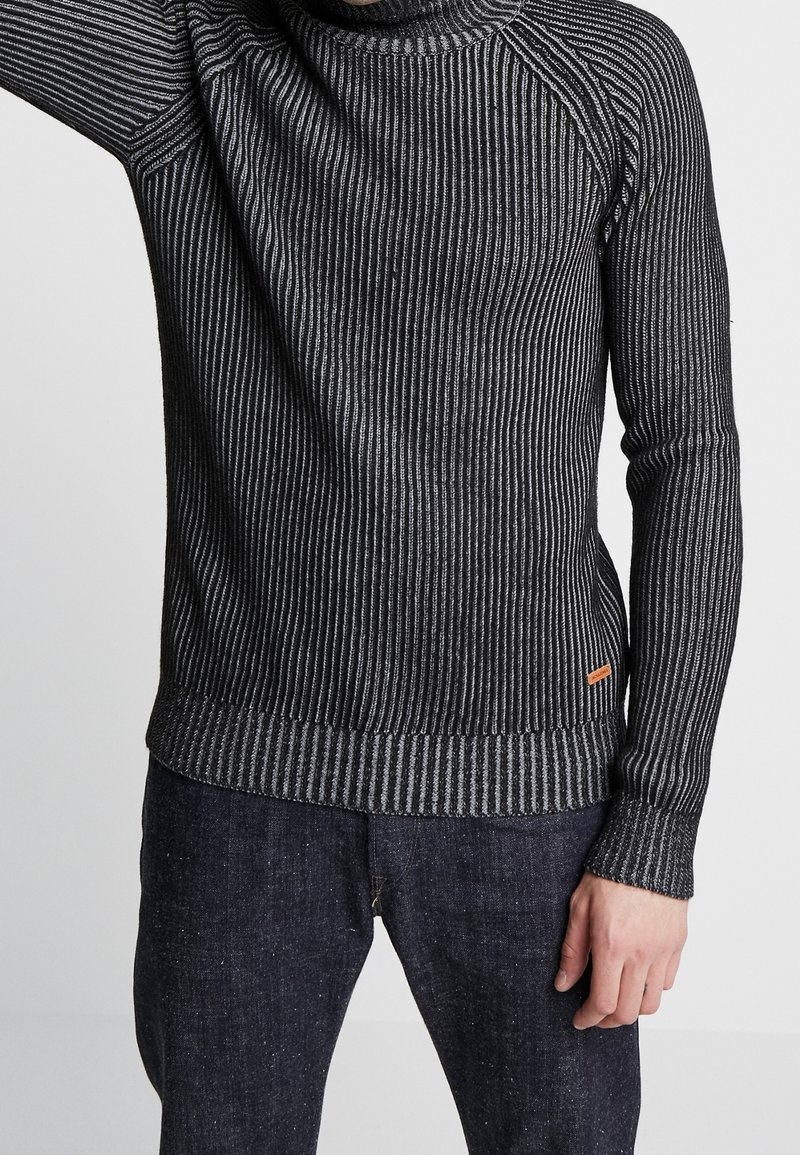 Svart och grå randig tröja med en ribbad textur, hög hals-design och en orange accenttagg i fållen. Långa ärmar med åtsittande mudd.