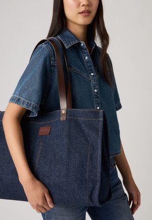 Donna con una camicia di jeans blu che porta una grande borsa a tracolla in denim con manici in pelle marrone sulla spalla.
