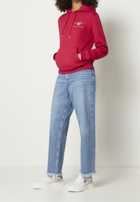 Czerwona bluza z kapturem z kieszenią z przodu i sznurkiem, zestawiona z jasnoniebieskimi jeansami o prostych nogawkach. Białe trampki z czerwonymi akcentami i postrzępionymi brzegami.
