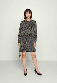 Vestido com estampado floral em preto com padrões verdes, vermelhos e roxos. Mangas longas, corte solto, comprimento acima do joelho, combinado com saltos pretos e uma bolsa pequena.