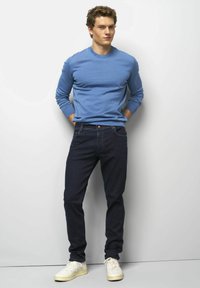 Long-sleeve blue shirt with ribbed cuffs, paired with dark jeans. The model wears white sneakers, with hands casually in pockets.