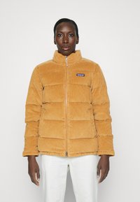 Chaqueta de plumas de pana en color amarillo mostaza, con un collar alto, cierre de cremallera y diseño acolchado. Presenta un parche con el logo en el pecho.