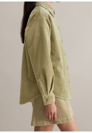 Person iført lysegrøn kordfløjls skjorte med knapper og matchende shorts, stående sidelæns mod en ensfarvet beige baggrund.