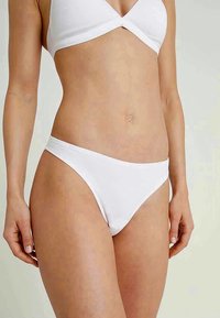 Woman wearing white bikini bottom and matching white bikini top, shown from mid-chest to upper thighs against plain background.