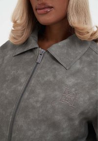 Giacca in finta suède grigia con zip frontale, ampio colletto e logo in rilievo discreto. Texture liscia con finitura opaca.