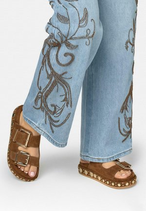 Sandalias de ante marrón con tiras de doble hebilla y suelas con plataforma tachonada, combinadas con jeans azul claro con diseños florales bordados en marrón.