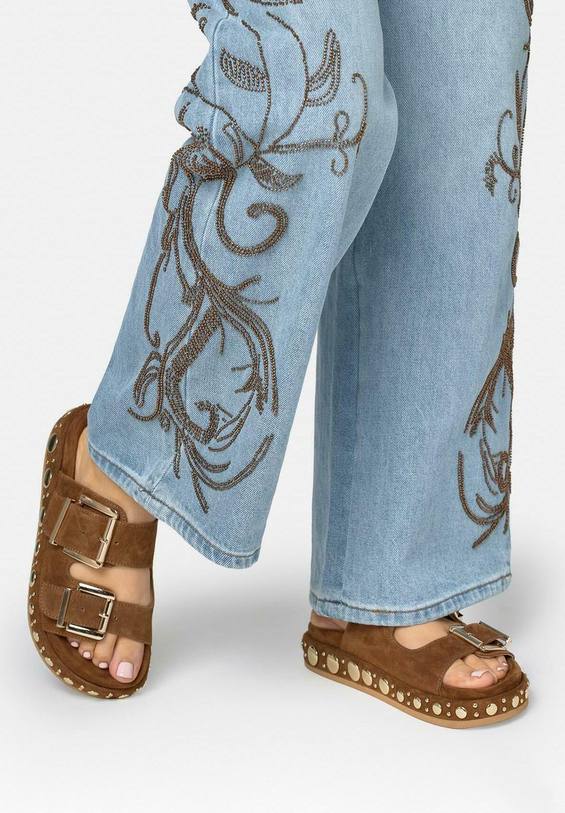 Sandales en daim marron avec brides à double boucle et semelles montantes cloutées portées avec un jean bleu clair orné de motifs floraux brodés marron.