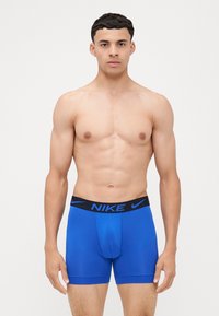 Modré boxerské trenky Nike s čiernym pásom obsahujúcim logo. Tkanina pôsobí hladko a tesne, pričom vystavuje moderný dizajn.
