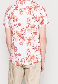 Camicia bianca a maniche corte con un motivo floreale in tonalità di rosa e arancione. Il retro mostra dettagli del design e un orlo leggermente curvo.