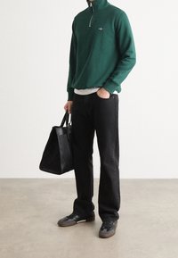 Zelený svetr na zip, šedé tričko pod ním, černé džíny, černá strukturovaná taška a tmavé boty; jednoduchý, funkční outfit.